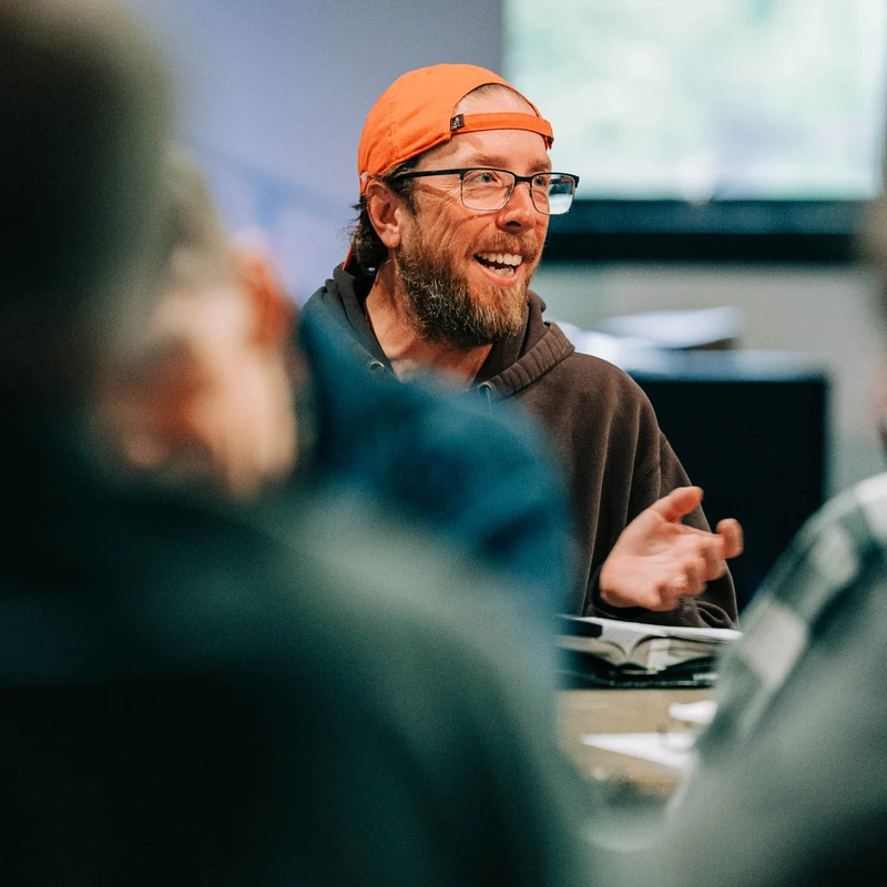 A man with a beard and glasses, wearing an orange baseball cap backwards and a hoodie, smiles and talks while sitting at a table in a group setting.