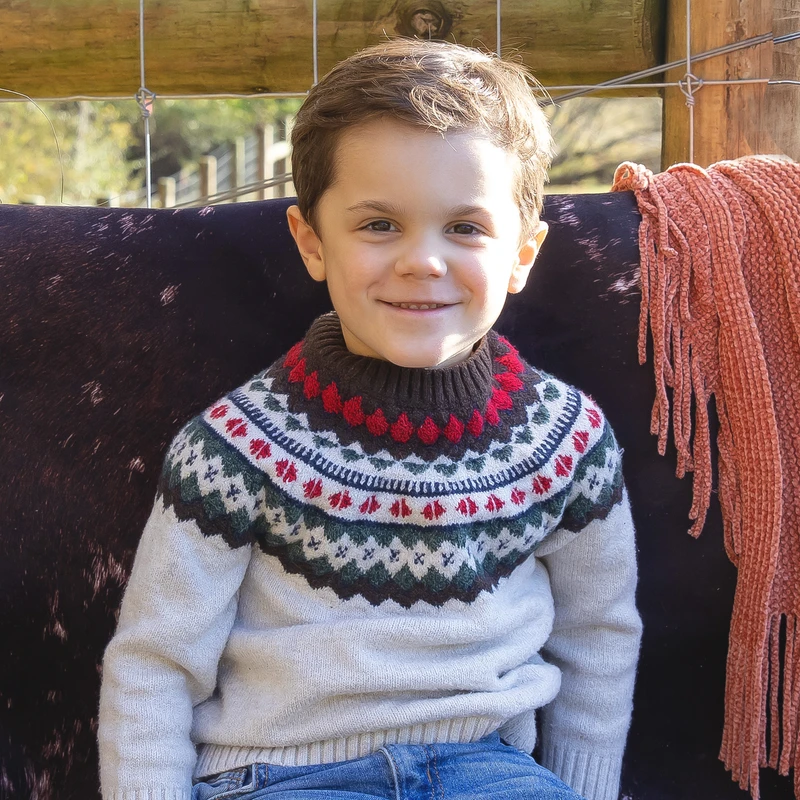 A young boy with brown hair smiles warmly while sitting outdoors. He is wearing a cozy, patterned knit sweater in red, green, and white.