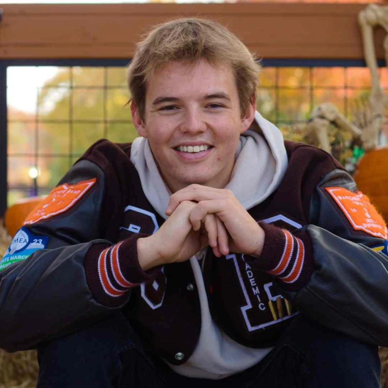 A happy young man with blondish-brown hair smiles while sitting outside, wearing a brown and black varsity jacket over a light-colored hoodie.