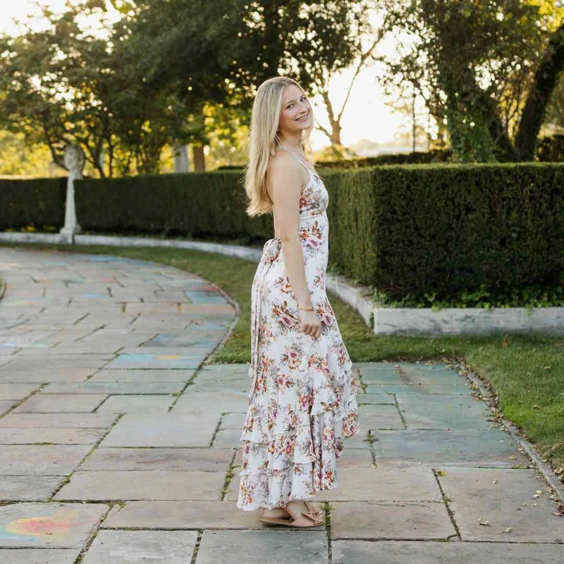 A young woman with long blonde hair smiles over her shoulder while standing on a stone path in a garden. She wears a long, white floral high-low dress.