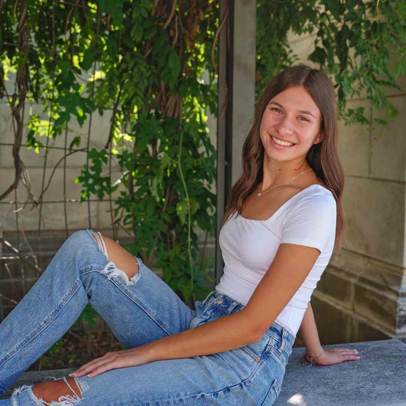 A young woman with long brown hair smiles while sitting on a stone bench. She wears a white top and ripped blue jeans, with lush green vines behind her.