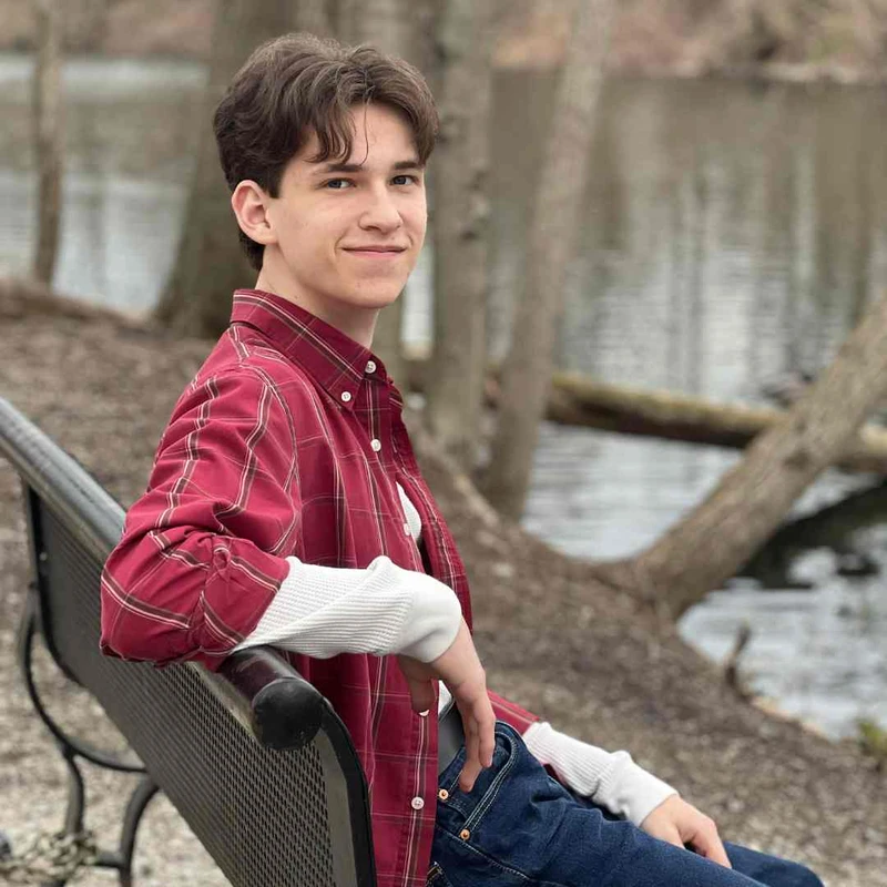 A young man with brown hair wearing a red plaid shirt sits on a black park bench, smiling gently at the camera. A body of water and trees are in the background.