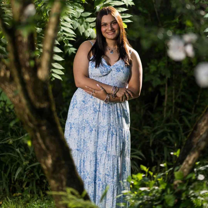 A young woman with long dark hair smiles while standing in a lush green garden. She wears a long blue and white floral dress and holds her arms crossed.