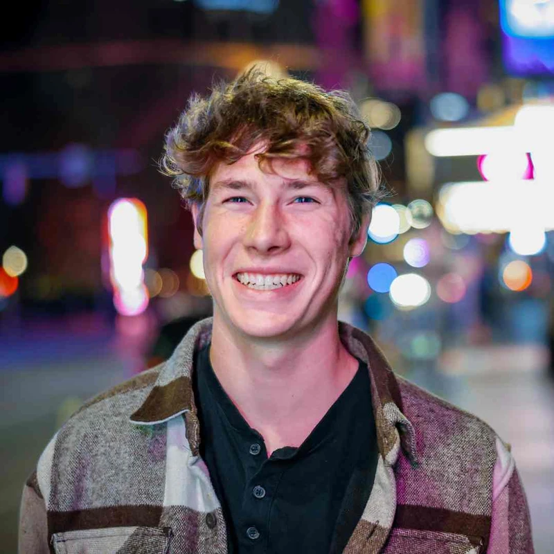 A headshot of a young man with curly light brown hair smiling at the camera. He is outdoors at night with a background of colorful, out-of-focus city lights.