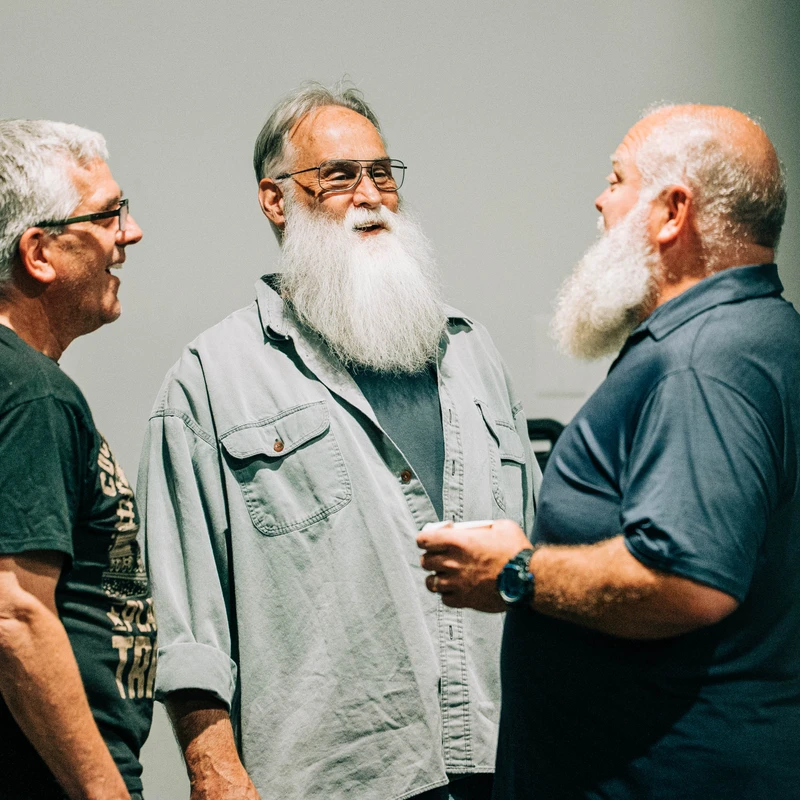 Three older men stand together indoors having a conversation. The two men on the right have long, full white beards and are smiling at each other.