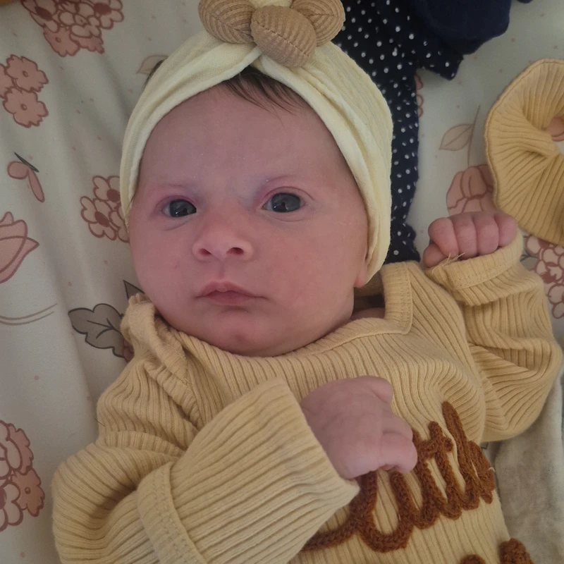 A newborn baby with dark, wide eyes looks up at the camera while wearing a yellow ribbed outfit and a matching knotted headband.