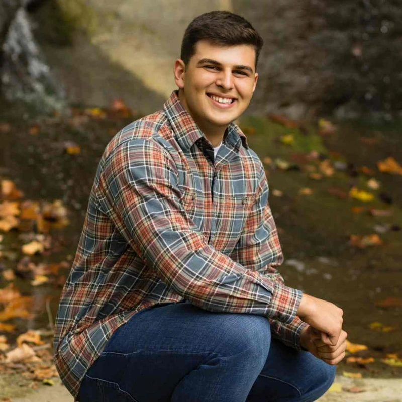 A young man with short dark hair smiles while crouching outdoors. He wears a plaid shirt and jeans, with a waterfall and autumn leaves in the background.