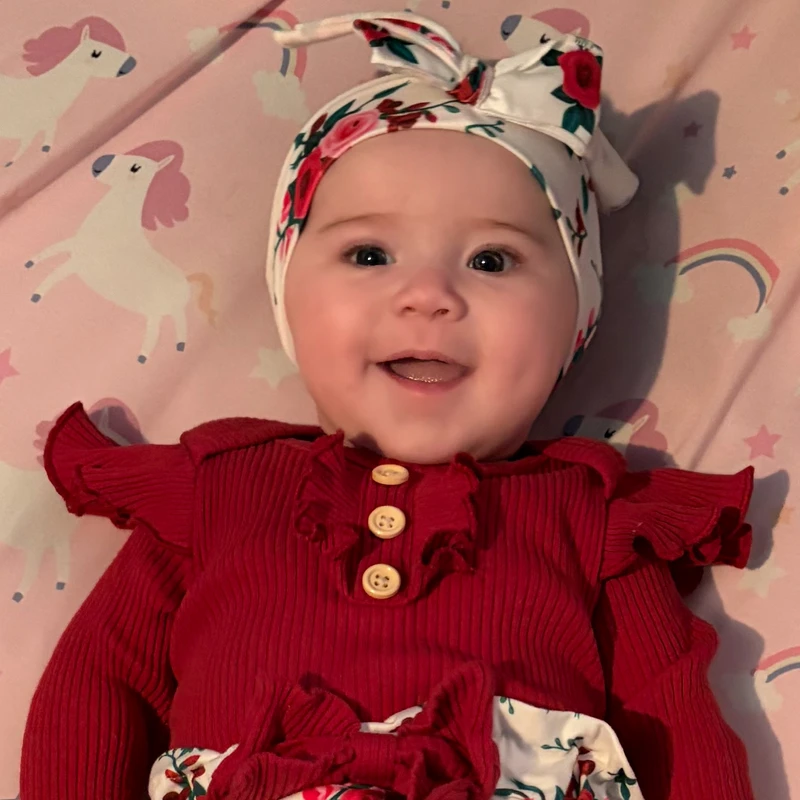 A smiling baby girl with dark eyes and chubby cheeks wears a red outfit and a floral headband, lying on a pink blanket with a unicorn and rainbow pattern.
