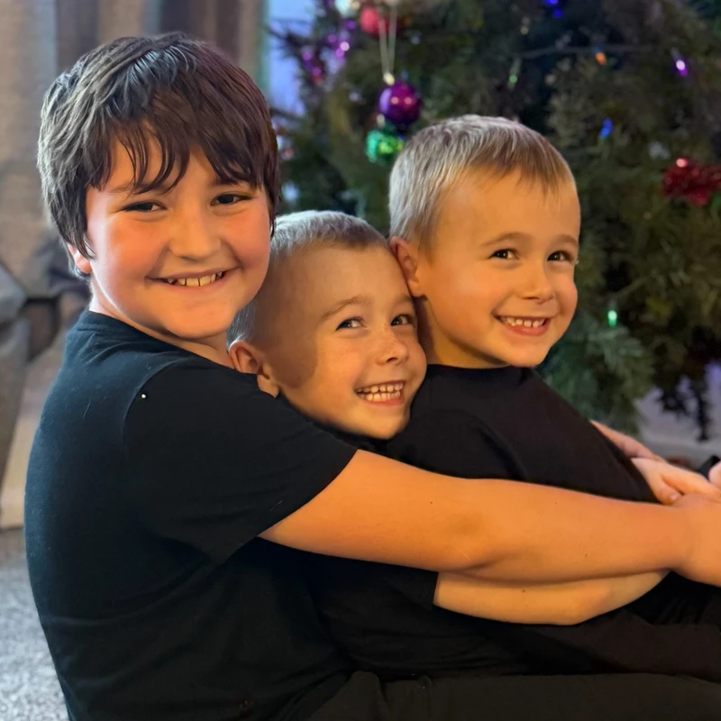 Three young brothers in matching black shirts smile and hug each other warmly in front of a decorated Christmas tree.