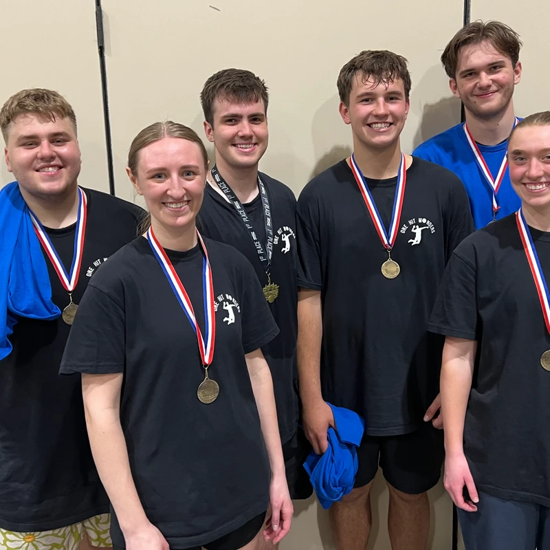 A group of six young adult volleyball players, wearing matching black t-shirts and gold medals, smile for a photo after a competition.