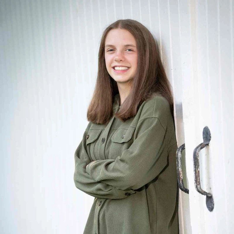 A happy teenage girl with long brown hair, wearing a green jacket, smiles while leaning against a white wall with her arms crossed.