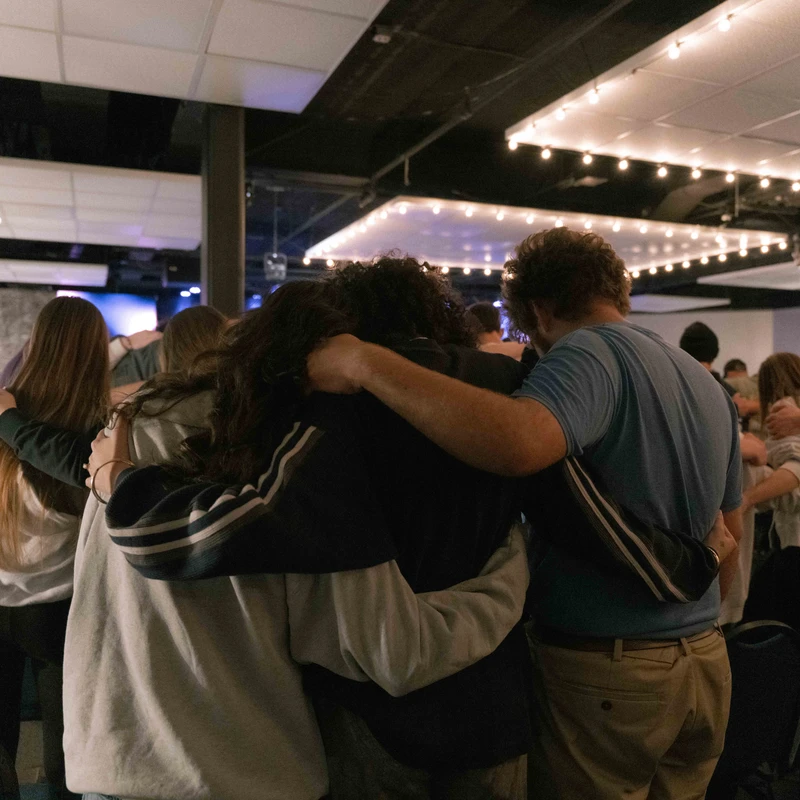 A group of people seen from behind, huddling in a circle with their arms around each other in a supportive gesture, likely praying in a large room.