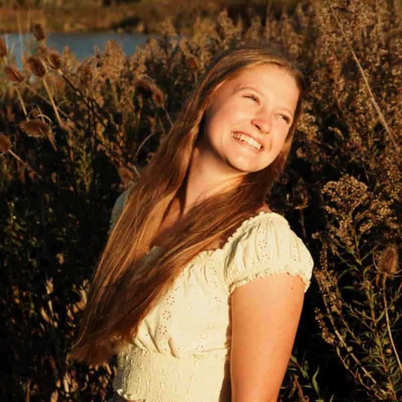 A young woman with long blonde hair smiles while standing in a field of tall, brown plants during golden hour. She wears a white top and looks happy.