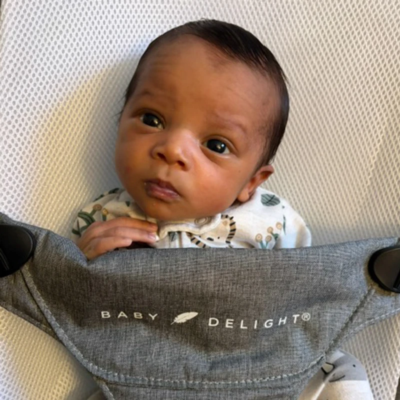 A close-up photo of a newborn baby with dark hair and eyes, looking alert while sitting in a grey bouncer seat with the words "Baby Delight" on it.