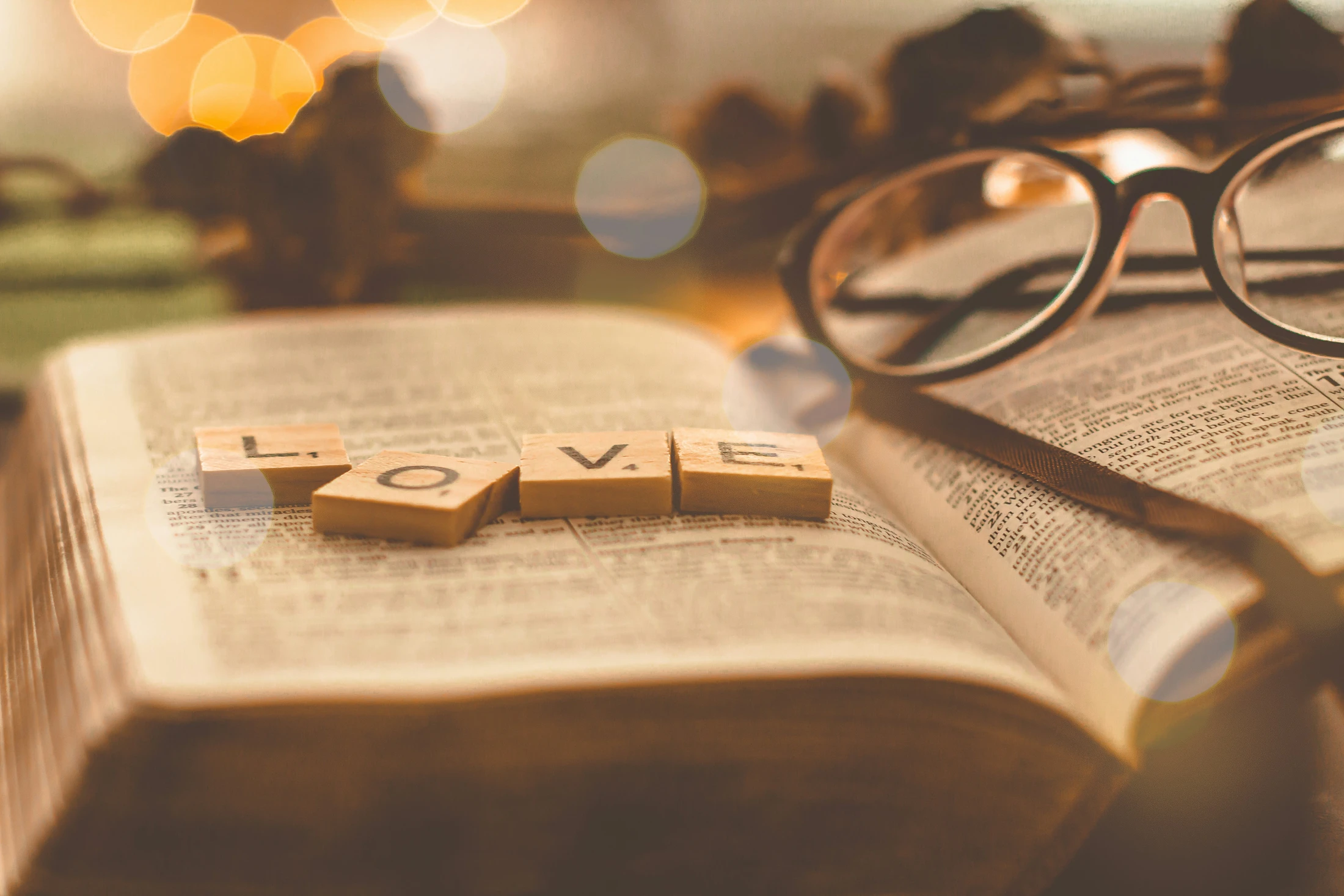 Wooden tiles spelling the word LOVE on the pages of an open book, possibly a Bible, with reading glasses nearby and soft, warm bokeh lights in the background.