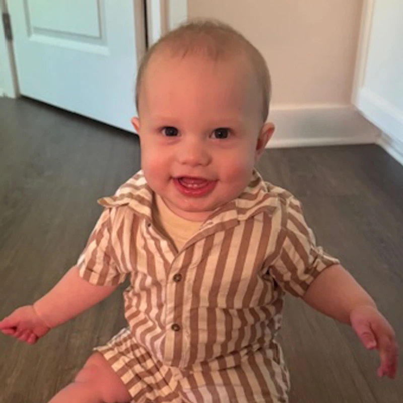 A smiling baby with light hair and blue eyes sits on a wood floor, wearing a tan and white striped one-piece outfit and looking at the camera.