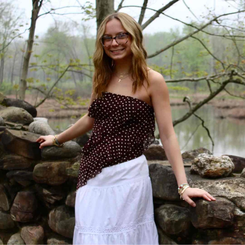 A smiling young woman with blonde hair and glasses leans on a stone wall. She wears a brown polka-dot top and white skirt, with a pond and trees behind her.