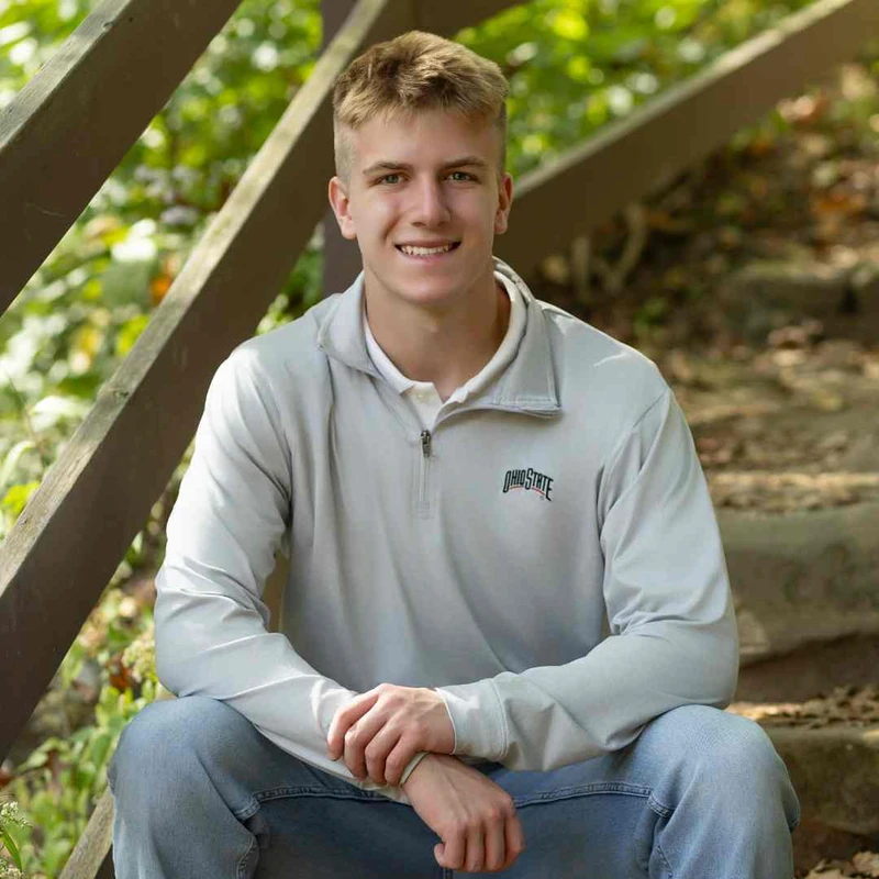 A smiling young man with short blond hair sits on outdoor steps. He wears a gray Ohio State pullover and jeans, with a leafy green background.