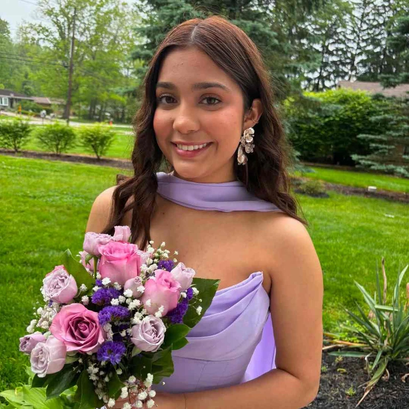 A smiling young woman with long brown hair, wearing a lavender gown and holding a bouquet of pink and purple flowers, stands outdoors on a green lawn.