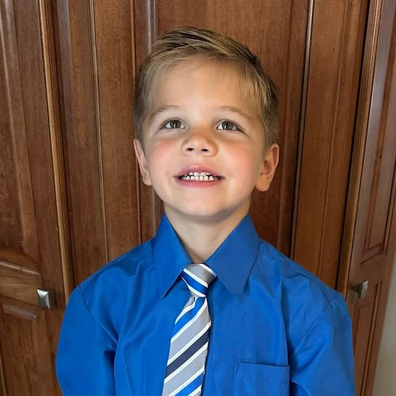 A smiling young boy with blond hair and brown eyes looks up. He is wearing a bright blue collared shirt and a striped tie in front of a wood-paneled door.