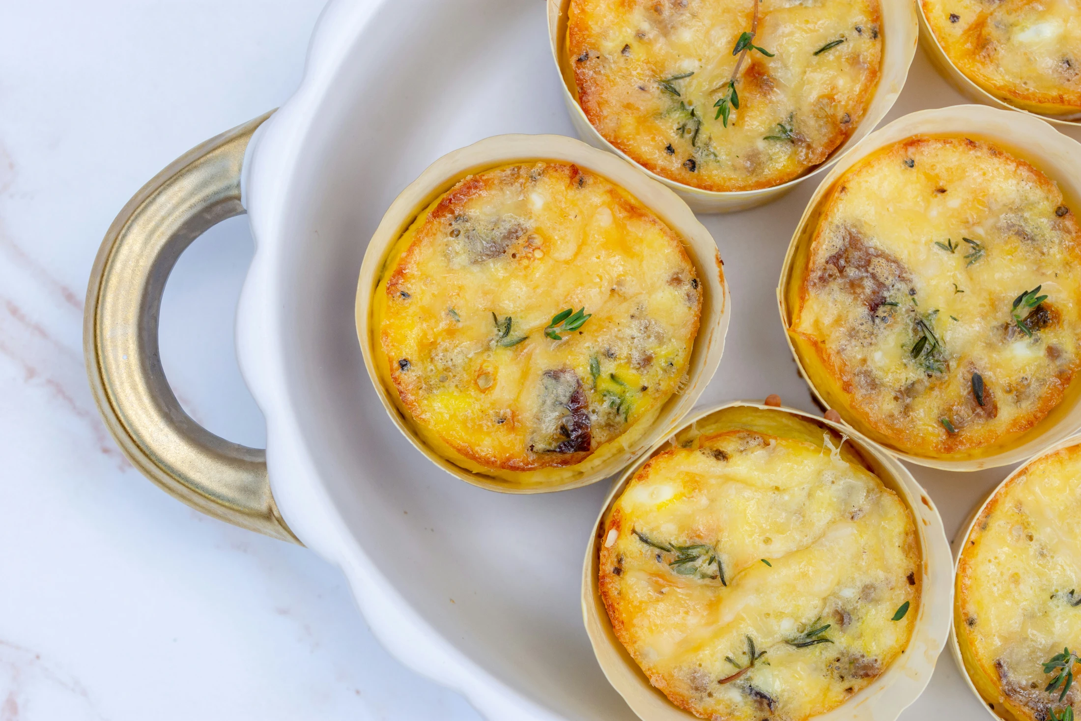 An overhead shot of individual savory egg bites, topped with fresh thyme, sitting inside a white ceramic baking dish with a decorative gold handle.