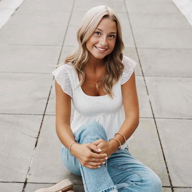 A young woman with blonde, wavy hair sits on a grey paved walkway, smiling at the camera. She is wearing a white blouse, blue jeans, and bracelets.