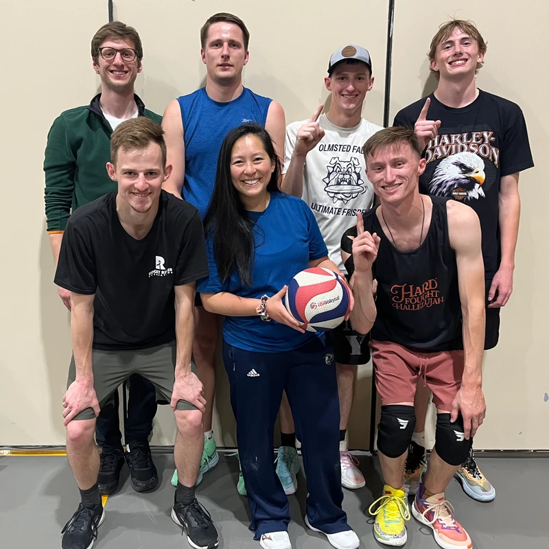 A group of seven men and one woman, a volleyball team, smile for a photo. The woman holds a volleyball, and several men hold up 'number one' fingers.