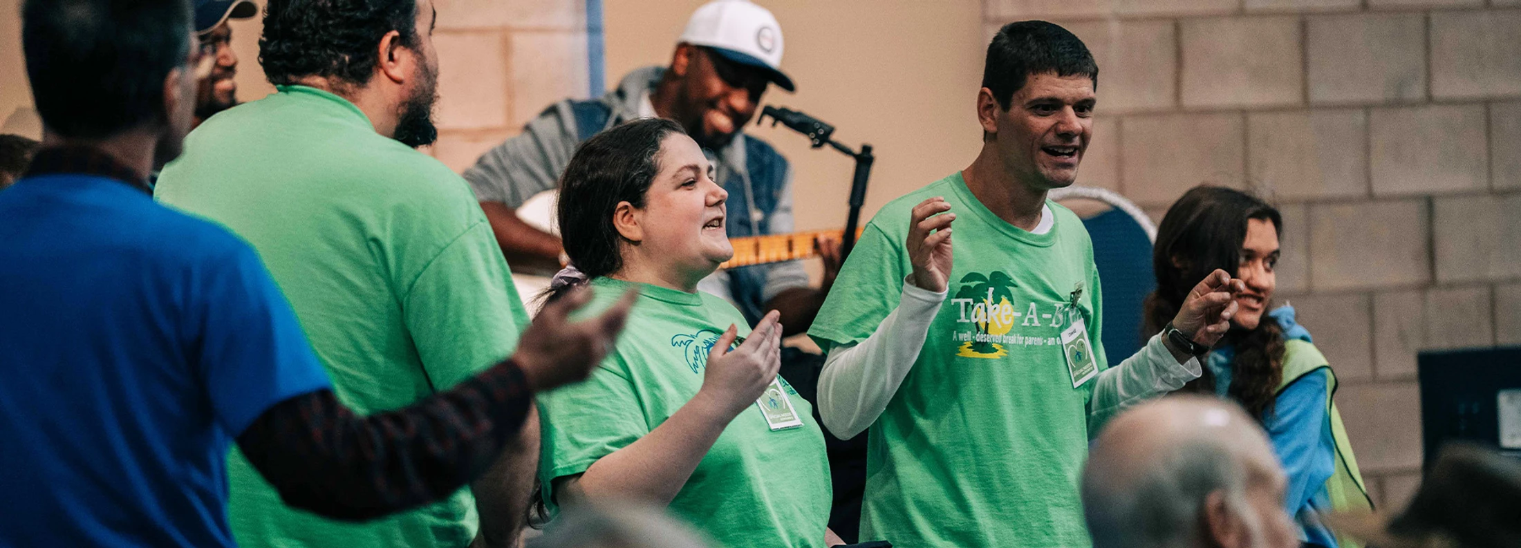A diverse group of smiling people in matching green t-shirts sing together with raised hands. A man plays a guitar in the background.