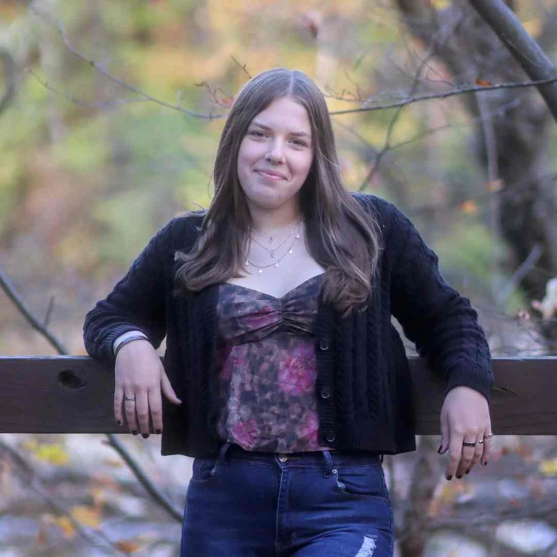A young woman with long brown hair smiles while leaning on a wooden fence. She wears a black cardigan, a floral top, and jeans in a wooded setting.