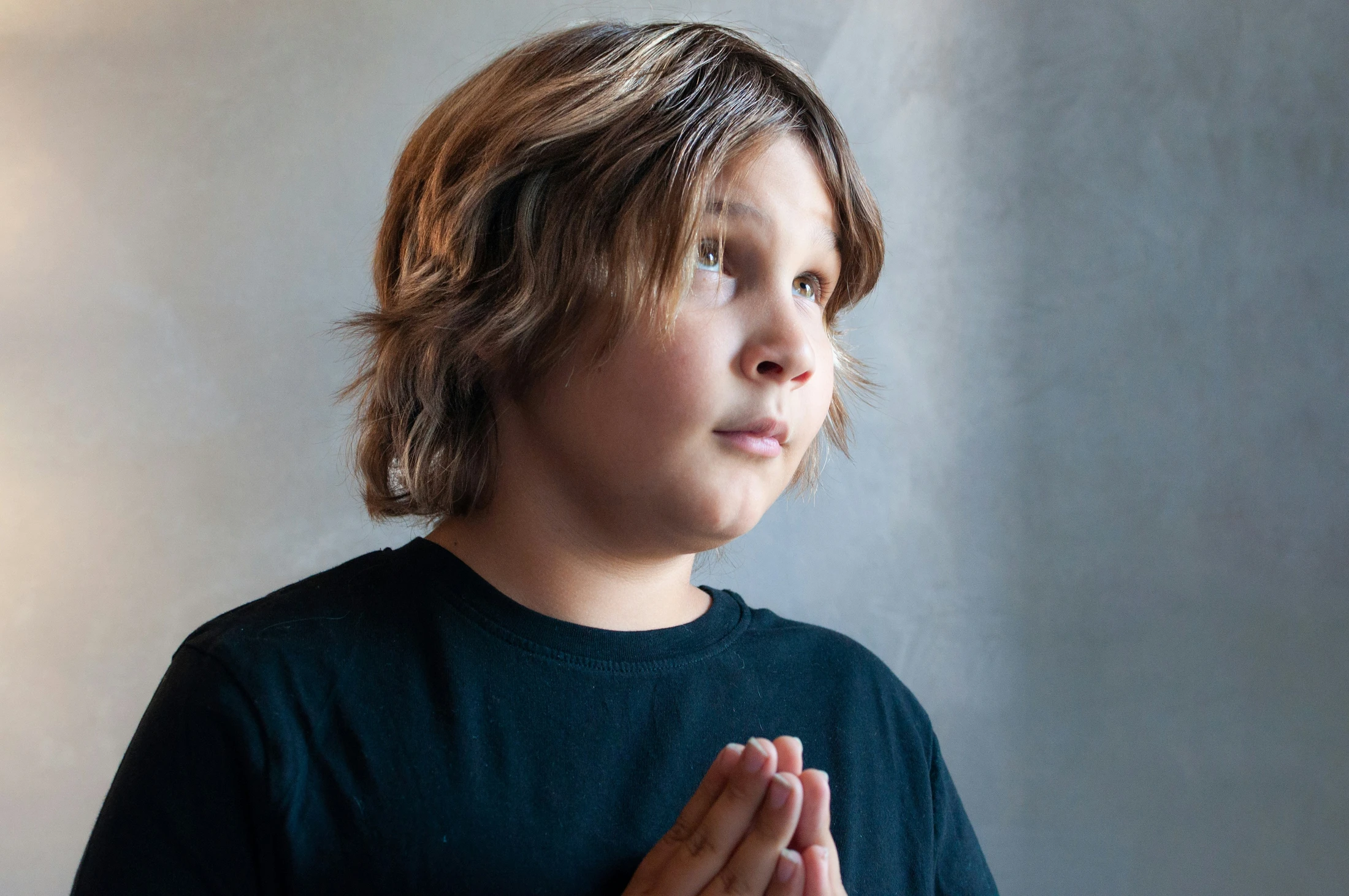 A young boy with shaggy brown hair and a black shirt looks up with a hopeful expression. His hands are clasped together in front of him as if in prayer.
