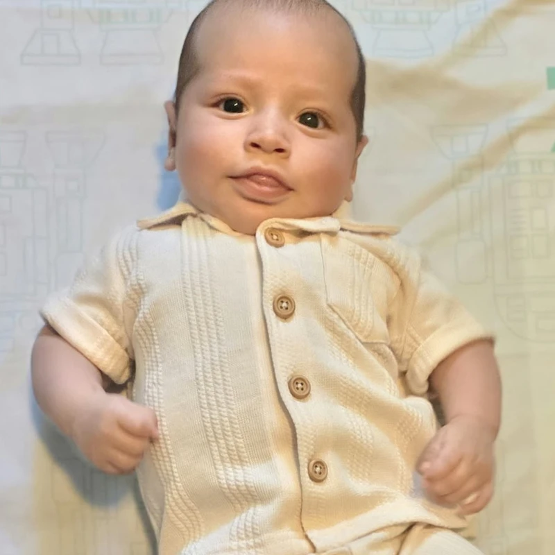 A baby with large, dark eyes lies on their back looking at the camera, wearing a cream-colored, textured, short-sleeved button-up shirt and matching pants.