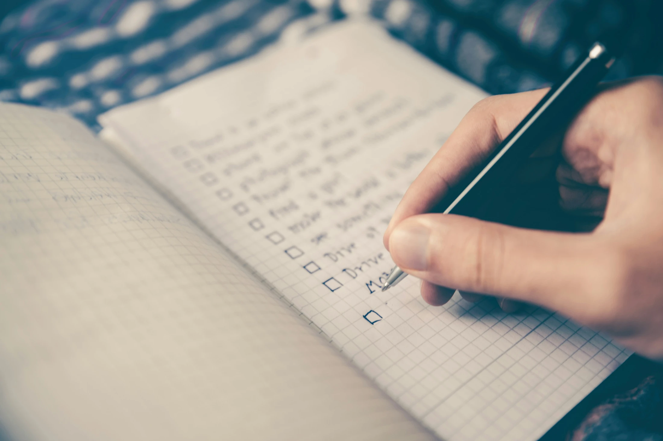 A close-up of a person's hand holding a black pen, writing a checklist with checkboxes on graph paper in an open notebook.