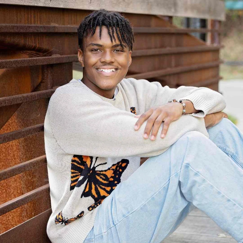 A young Black man with twisted hair smiles warmly while sitting outdoors. He is wearing a cream sweater with an orange and black butterfly design.