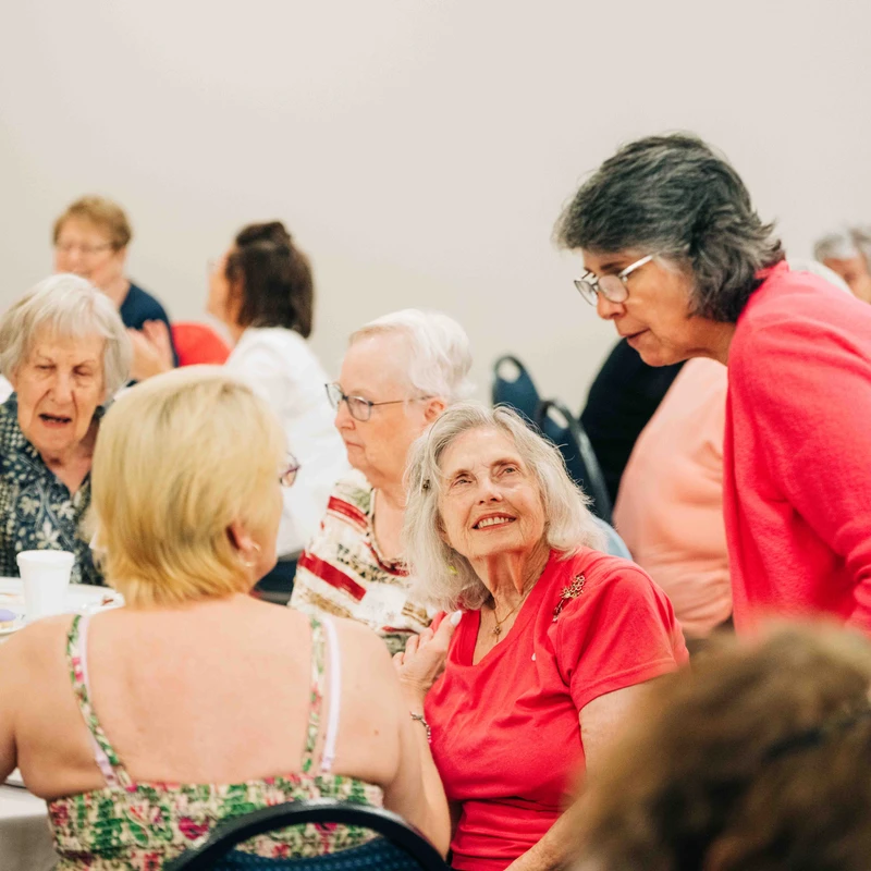 A group of older women sit and stand around a table indoors, talking. A woman in a red shirt smiles as she looks up at another woman leaning in to speak.