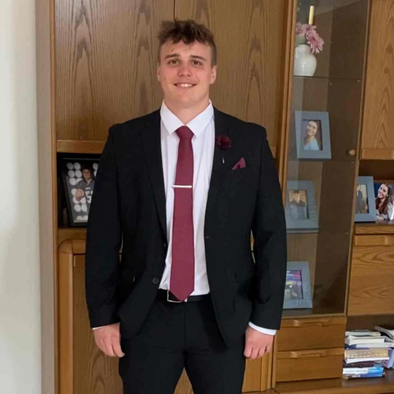 A smiling young man in a black suit, white shirt, and burgundy tie stands in front of a wooden display cabinet in a home.