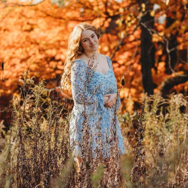 A young woman with long wavy hair, wearing a blue floral dress, stands among tall dry grasses in a field with vibrant orange autumn trees in the background.