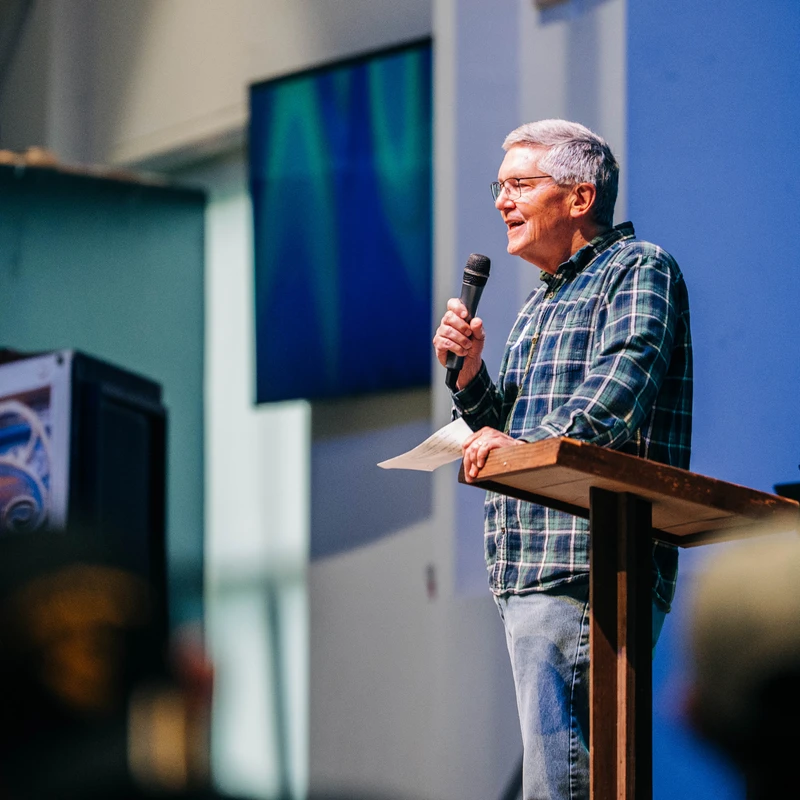 An older man with glasses and a plaid shirt stands at a wooden lectern on a stage, speaking into a microphone with a blue background.