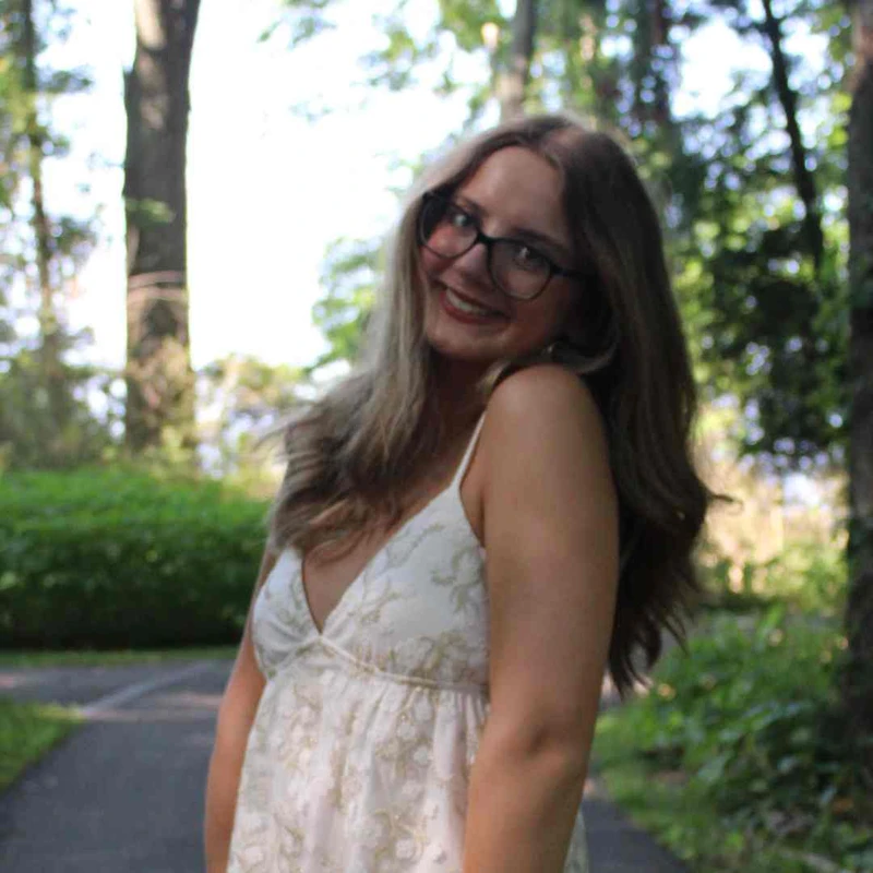 A young woman with long brown hair and glasses smiles at the camera. She is wearing a white summer dress and standing on a path in a wooded park.