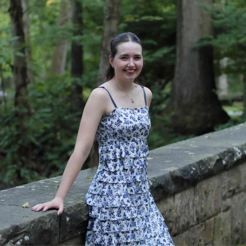 A smiling young woman with dark hair, wearing a tiered blue and white floral sundress, leans against a stone wall in a forest setting.