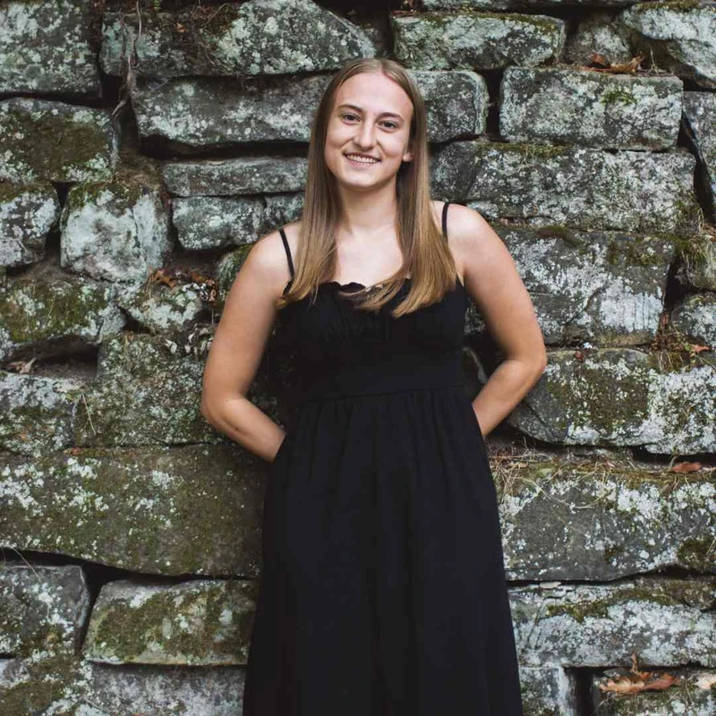 A young woman with long blonde hair, wearing a black dress, smiles while leaning against a rustic, moss-covered stone wall.