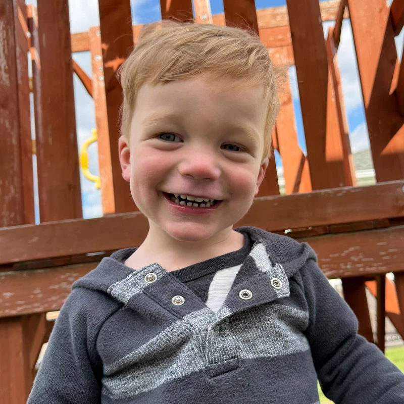 A close-up of a smiling young boy with strawberry-blonde hair, wearing a grey striped hoodie, sitting on a wooden playset on a sunny day.