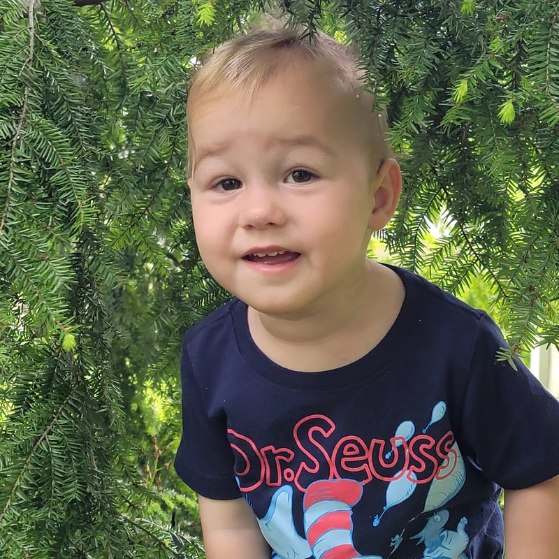 A young toddler with blond hair and a blue Dr. Seuss t-shirt smiles at the camera while standing in front of a lush green evergreen tree.