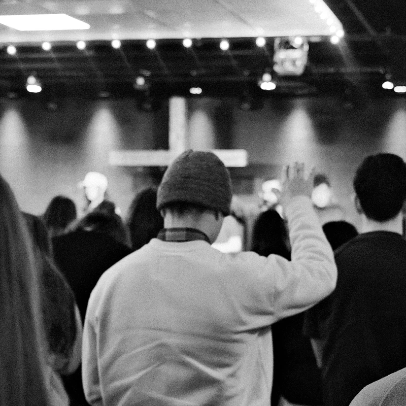 A grainy, black and white photo from behind a crowd looking toward a stage with a cross. A person in a beanie raises their hand in an act of worship.
