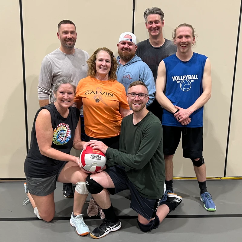 A group of seven adult volleyball players, five men and two women, smile for a team photo in a gym. One man in front kneels and holds a volleyball.