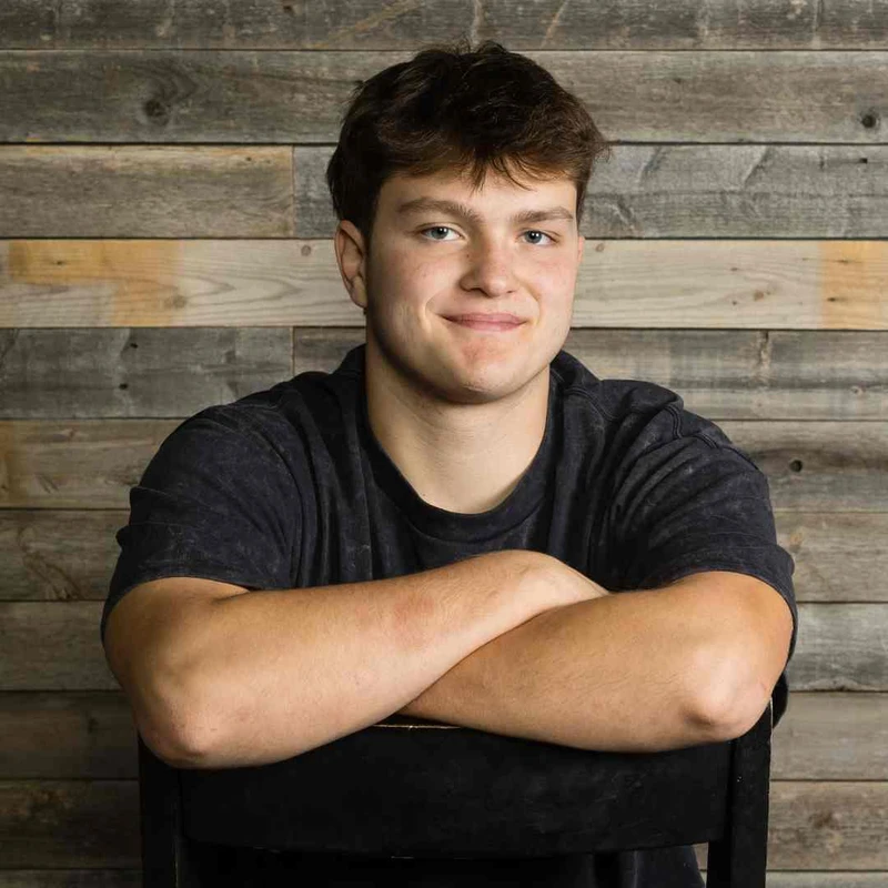 A headshot of a young man with short brown hair and blue eyes smiling gently. He is sitting on a chair with arms crossed, in front of a wooden plank background.