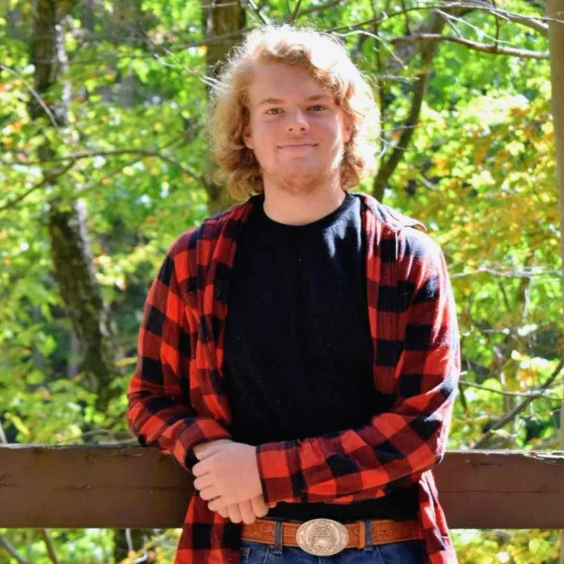 A young man with curly blonde hair smiles at the camera. He wears a red and black plaid shirt over a black t-shirt and leans on a railing in a forest.