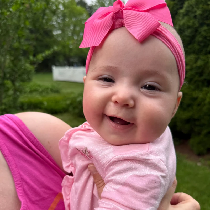 A close-up of a happy baby girl smiling at the camera. She is wearing a pink outfit and a large, bright pink bow headband and is being held outdoors.