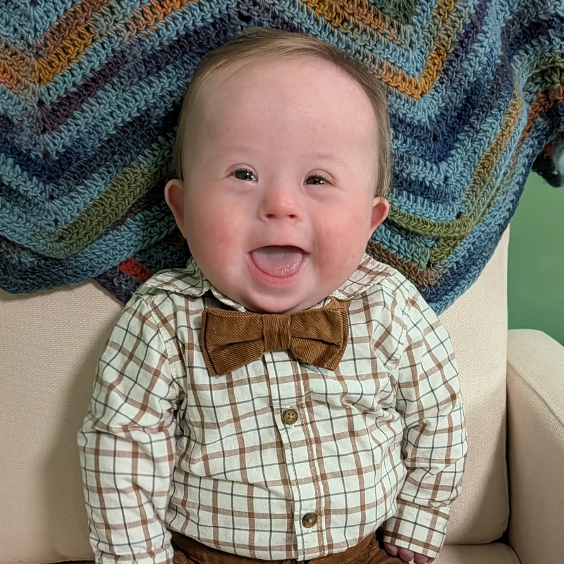 A happy baby boy with Down syndrome, dressed in a plaid shirt and brown bow tie, smiles wide for the camera while sitting with a colorful crocheted blanket.