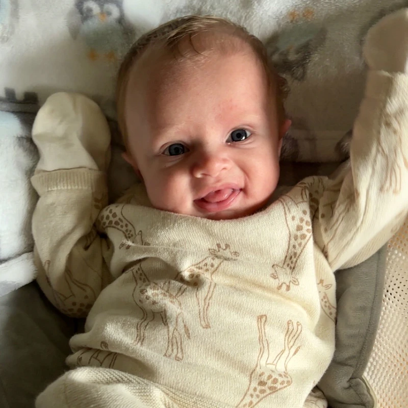 A smiling baby with blue eyes lies on a blanket, sticking their tongue out. The baby is wearing a cream-colored onesie with a giraffe pattern.