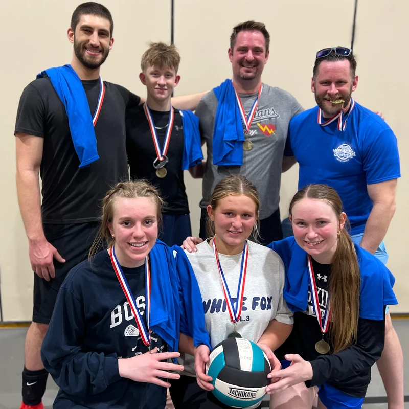 A smiling co-ed volleyball team of seven people poses for a photo in a gym. They are all wearing medals, and the three girls in front hold a volleyball.